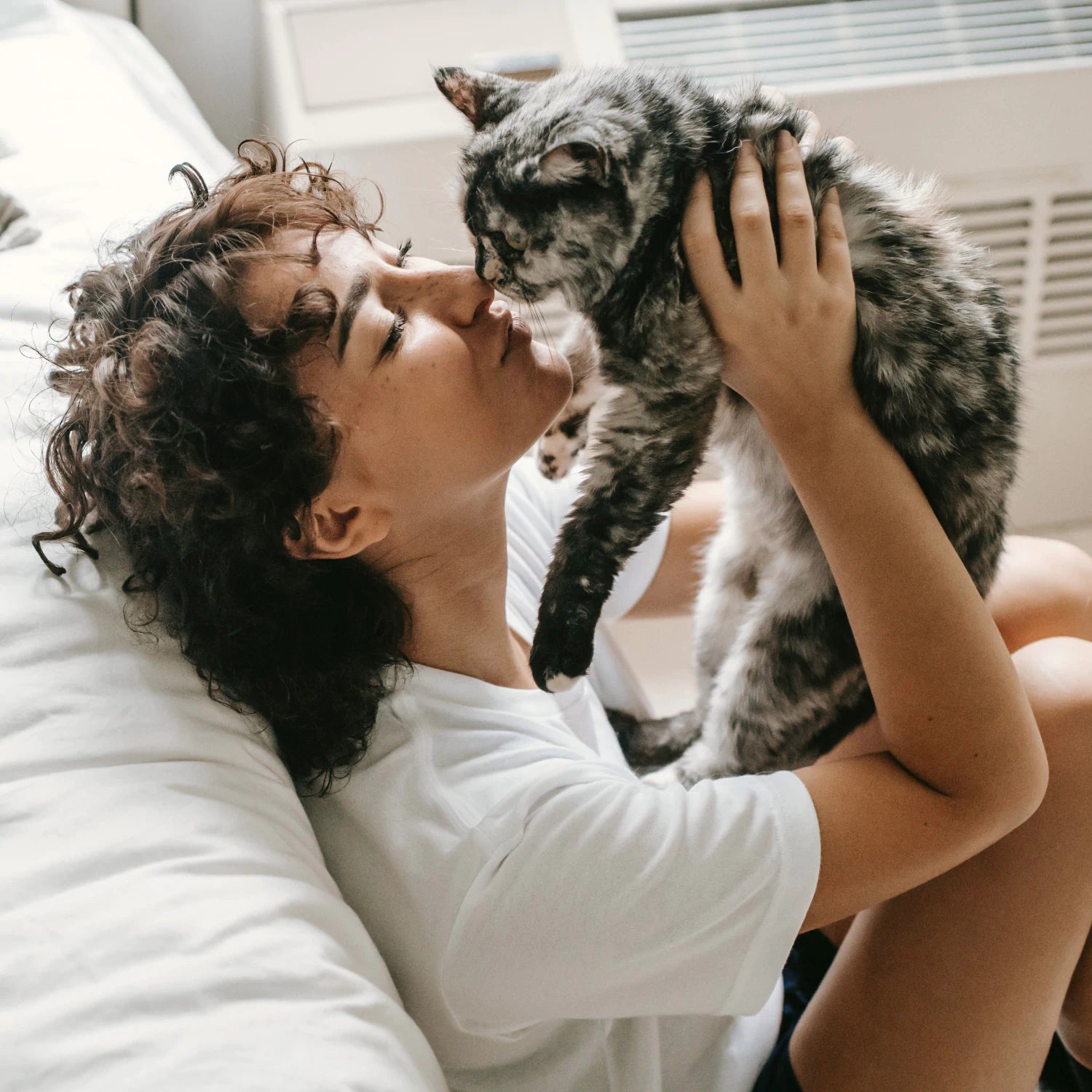Person sitting on the floor next to a bed in a bright room, holding a grey and black cat to their face.