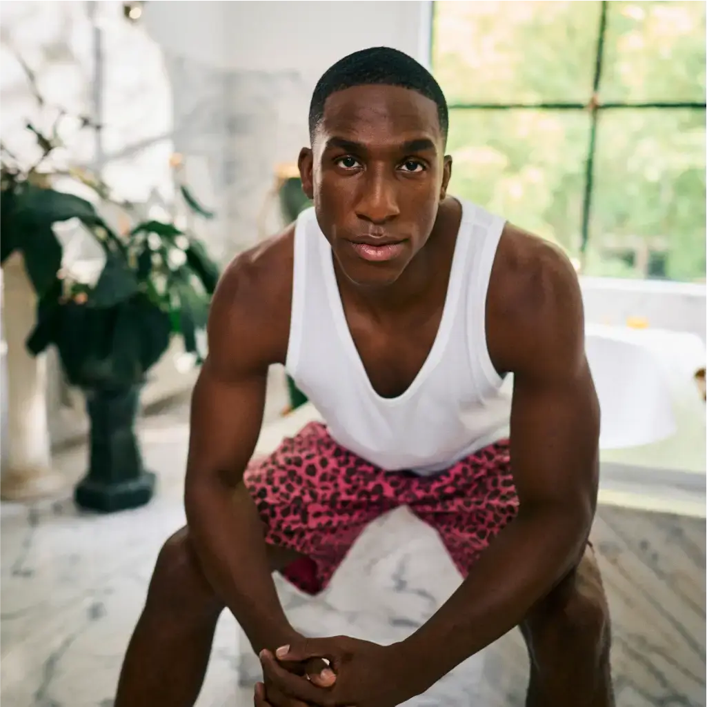 A man in a white tank top and pink leopard-print shorts sits with folded hands on an indoor marble floor. Large windows and plants are visible in the background.