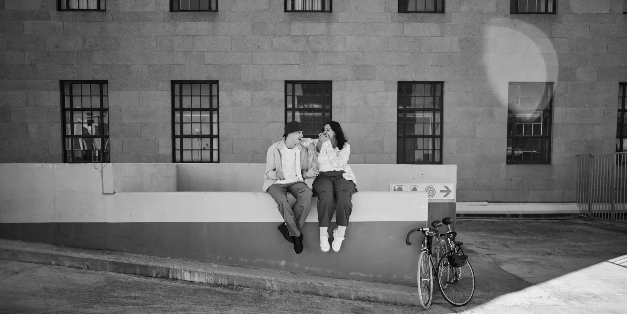 In front of a building with multiple windows, two people are sitting on a low wall, with a bicycle standing next to them.