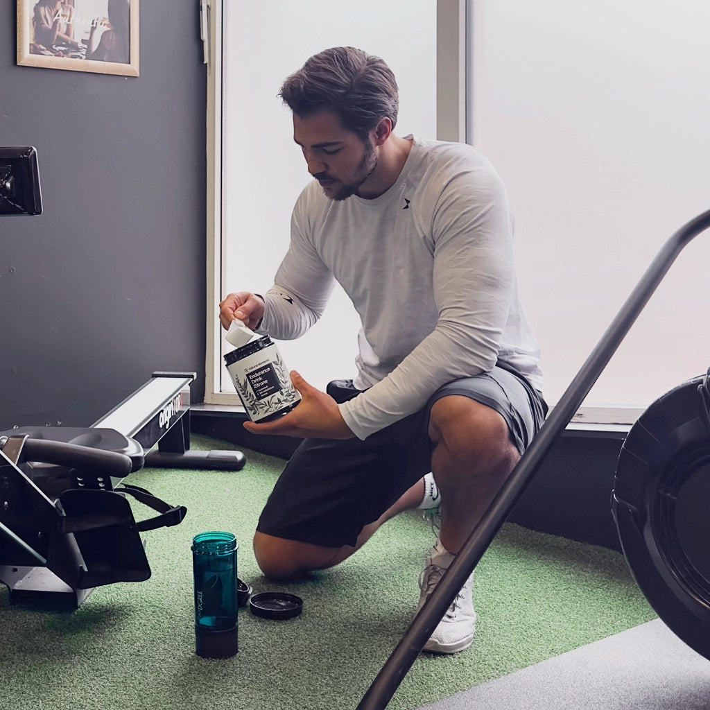 A man kneels on the gym floor, scooping powder from a container into a shaker bottle next to a rowing machine.