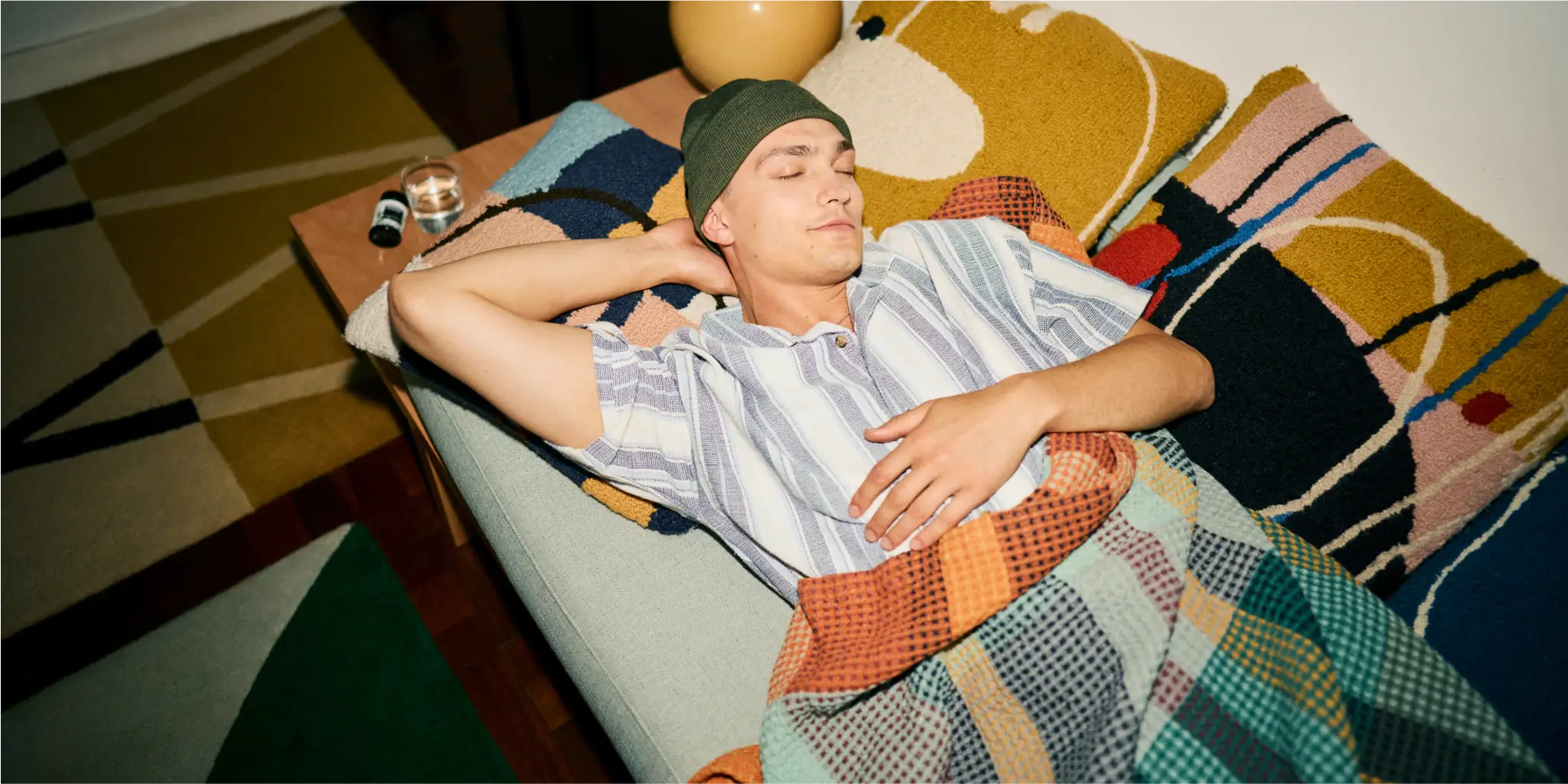 A person in a striped shirt and beanie lies on a sofa, covered with a colorful blanket, with eyes closed and an arm resting behind their head.