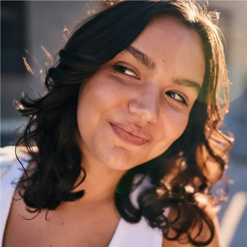A person with wavy, dark hair and a white top smiles slightly while looking to the side in the sunlight.