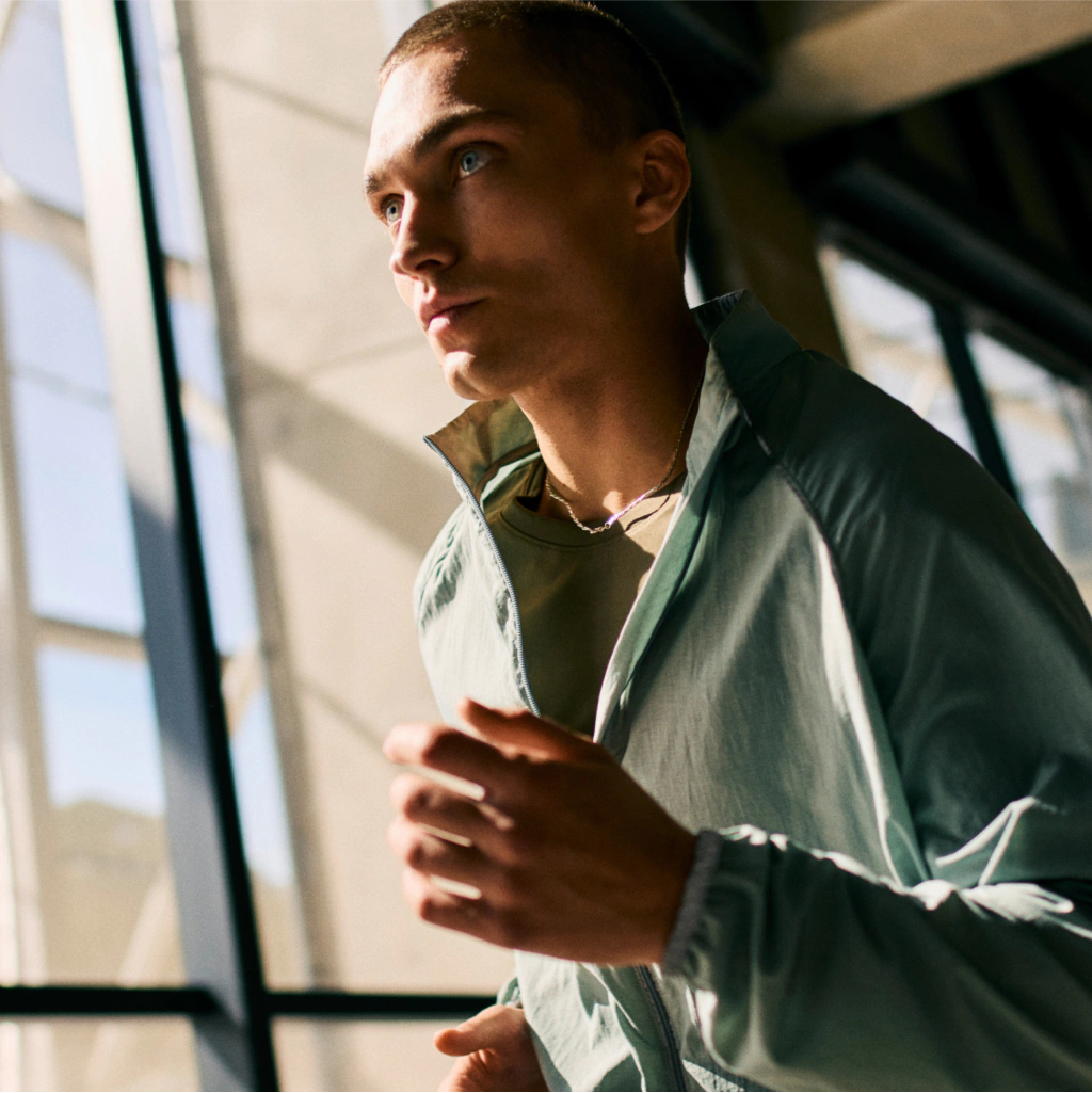 A person in a light jacket is jogging indoors near large windows, with sunlight streaming in from outside.