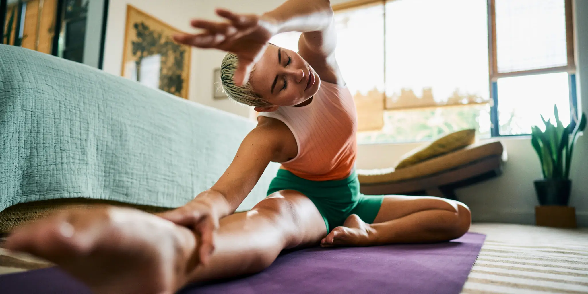 Person sitting on a yoga mat in a living room, stretching an arm overhead and reaching towards their extended leg.