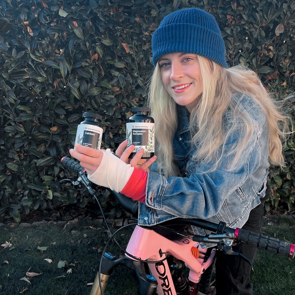 A blonde woman balances two bottles on her bike, surrounded by natural elements.